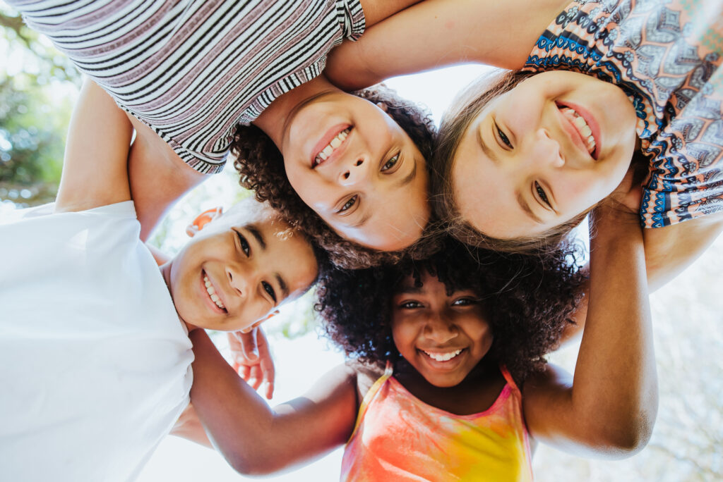 4 joyful children holding each other in a circle and smiling down at a camera.