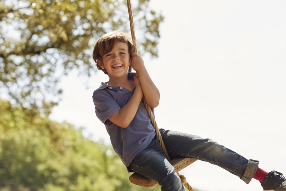 Portrait of cheerful boy playing on swing. Happy male is wearing casuals. He is enjoying against clear sky.